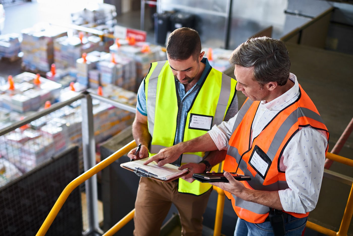 Two men in neon safety vests stand in a warehouse and consult a document on a clipboard.